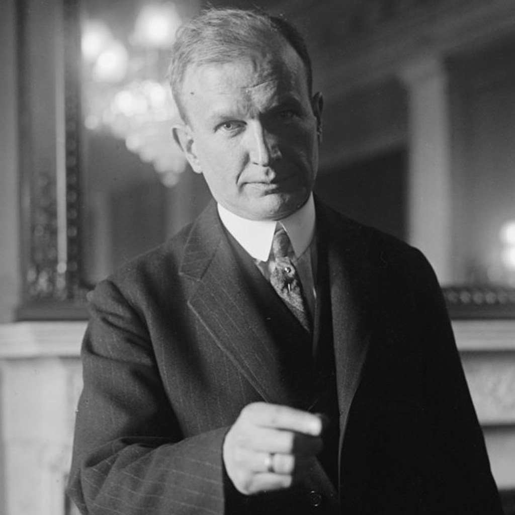 A serious man in a formal suit gestures with his right hand while looking directly at the camera, with a blurred chandelier in the background.